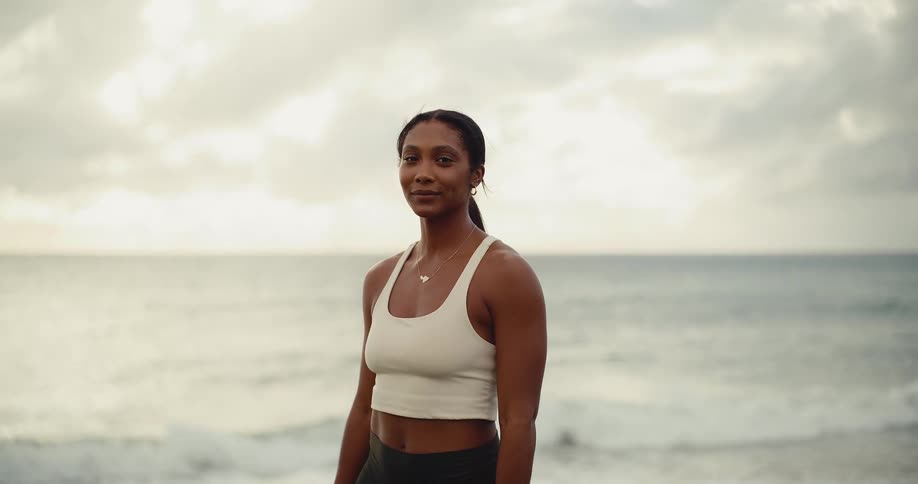 A calm and confident woman in athletic wear opens her eyes and smiles while standing by the ocean at sunrise