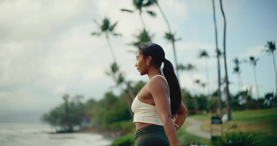 A fit woman in athletic clothing stretches her leg while standing in a scenic tropical park by the ocean