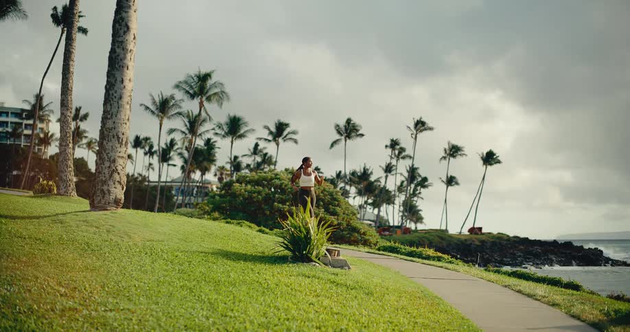 Slow motion shot of a beautiful athletic woman running along a coastal path with palm trees and the ocean