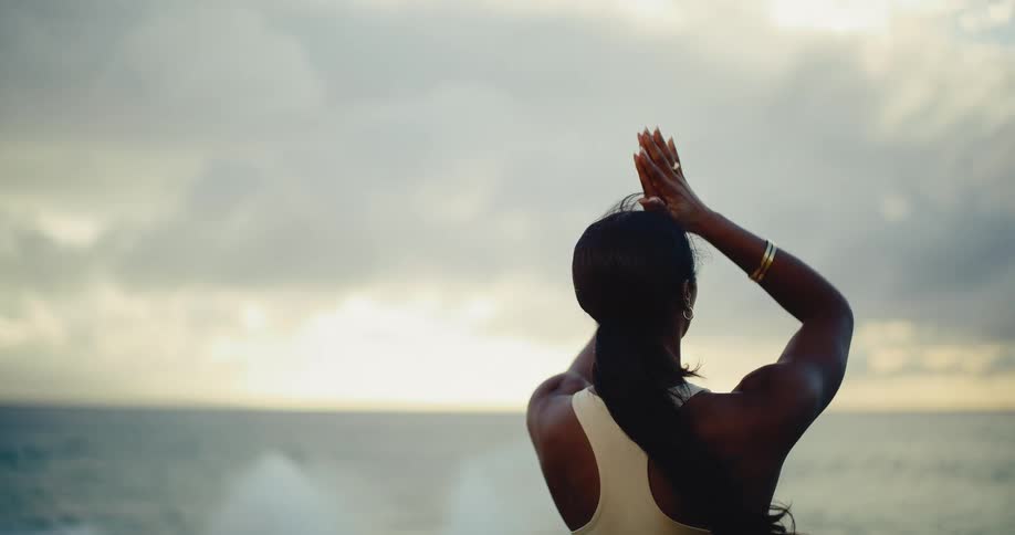From behind, a fit woman in athletic wear practices a slow yoga flow, raising her arms to the sky while overlooking a tranquil ocean at sunrise