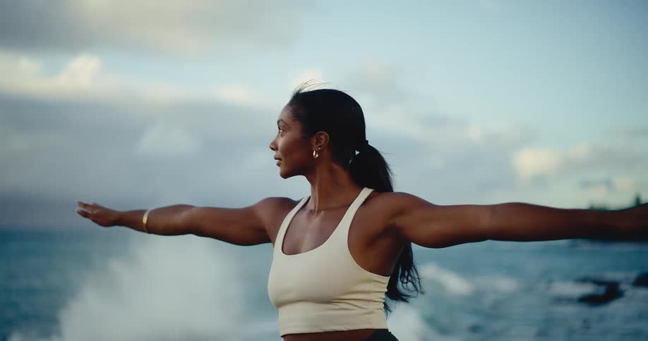 A strong and graceful woman transitions into a yoga warrior pose in slow motion with the ocean waves crashing behind her