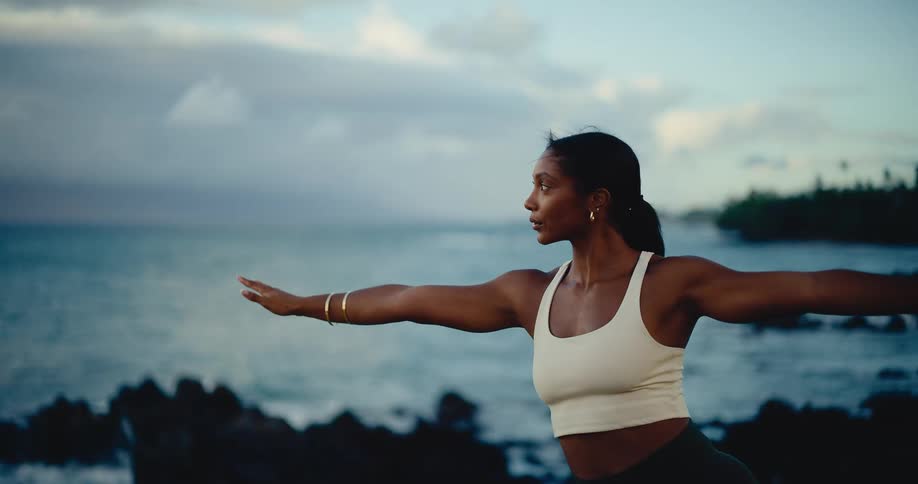 A focused woman gracefully practices a yoga flow in slow motion on a rocky coast with the ocean behind her