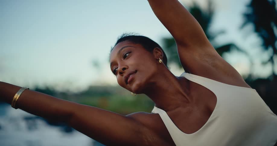 Close up slow motion of a beautiful woman of color practicing a serene yoga stretch by the ocean at dusk