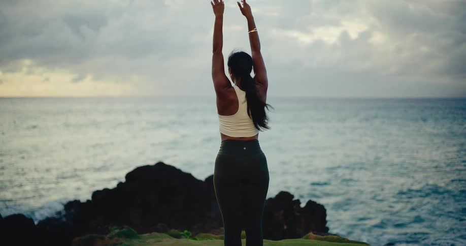 A fit woman in athletic wear practices a peaceful yoga flow on a grassy cliff overlooking the ocean at dawn in slow motion