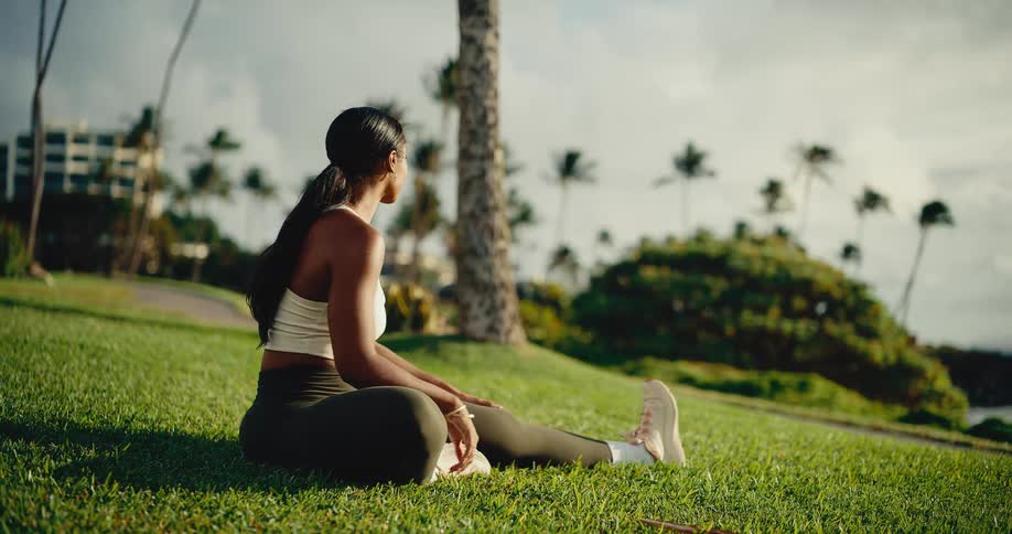 A fit woman performs a seated leg stretch on a grassy lawn by the ocean in a tropical park at sunrise