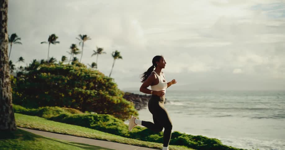 Beautiful athletic woman running along a coastal path lined with palm trees at sunrise in slow motion