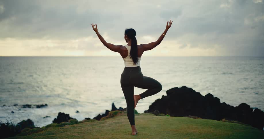 A fit woman with her back to the camera holds a yoga tree pose on a grassy cliff overlooking the ocean at dusk