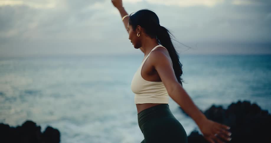 A fit and focused woman gracefully flows through yoga poses on a rocky shore with the ocean in the background at dawn