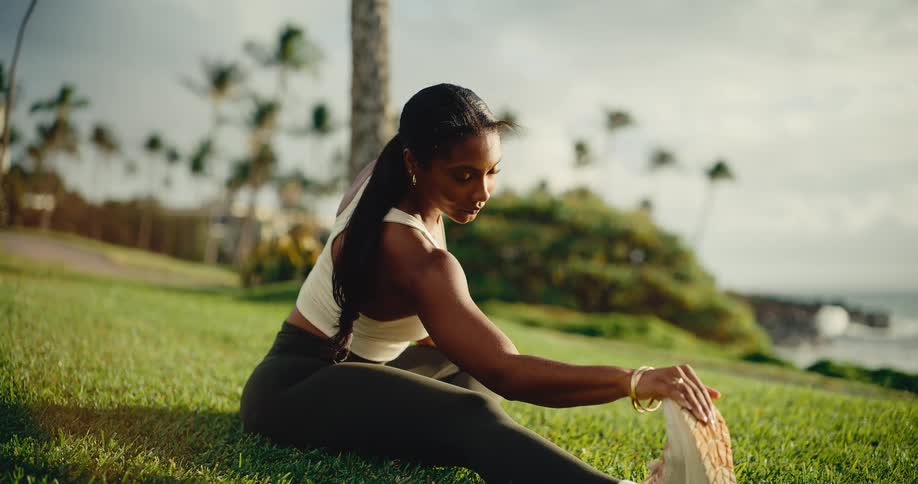 A beautiful athletic woman performs a deep stretch on a lush green lawn overlooking the ocean in a tropical paradise at sunrise