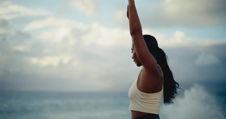 A woman in athletic wear mindfully performs a yoga flow in slow motion, stretching her arms towards the expansive ocean and cloudy sky