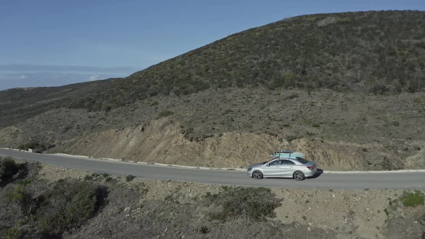 An aerial view shows a silver sedan with surfboards on the roof driving along a scenic coastal highway through rolling hills on a sunny day