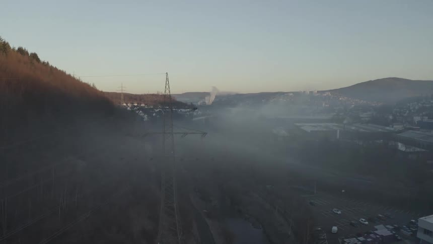 An aerial view ascends over a large transmission tower in a misty valley with an industrial town in the background at sunrise