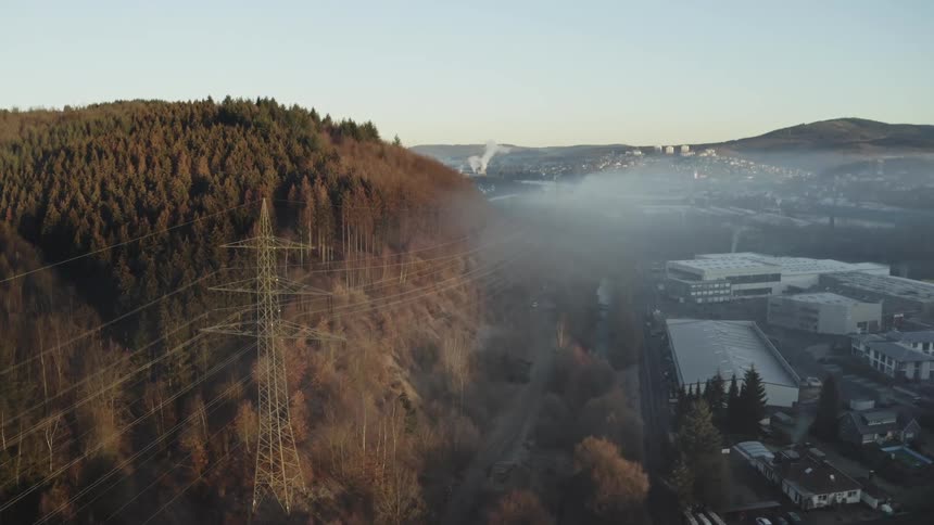 Flying past a transmission tower and power lines that separate a sunlit forest from a foggy industrial valley at sunrise