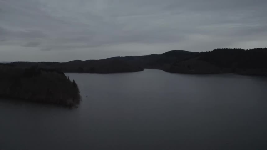 A still aerial shot captures a dark, expansive lake nestled among rolling, forested hills under a somber, overcast sky at twilight