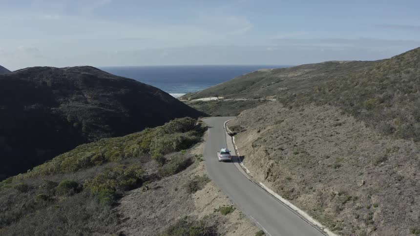 Aerial shot of a car with a surfboard driving on a winding coastal road through the mountains towards the sea