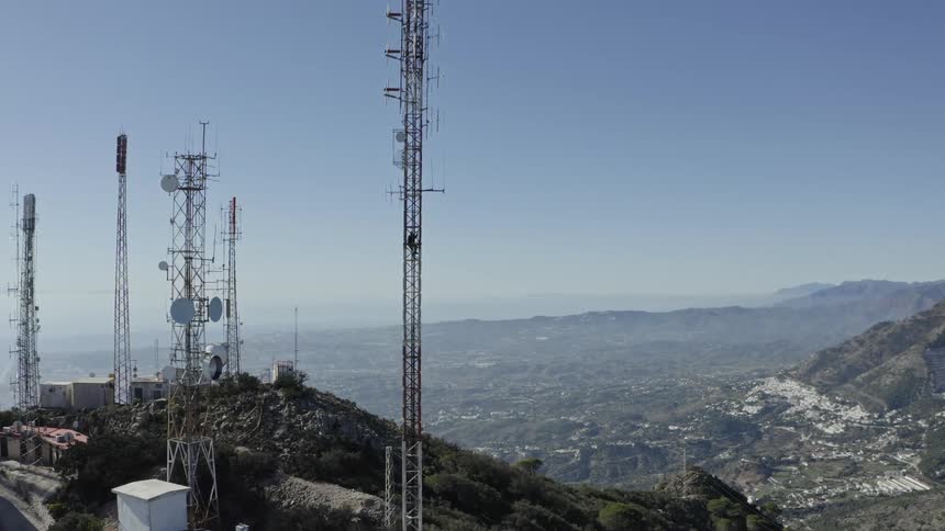 A worker climbs a tall telecommunication tower on a mountaintop overlooking a sprawling valley on a clear day