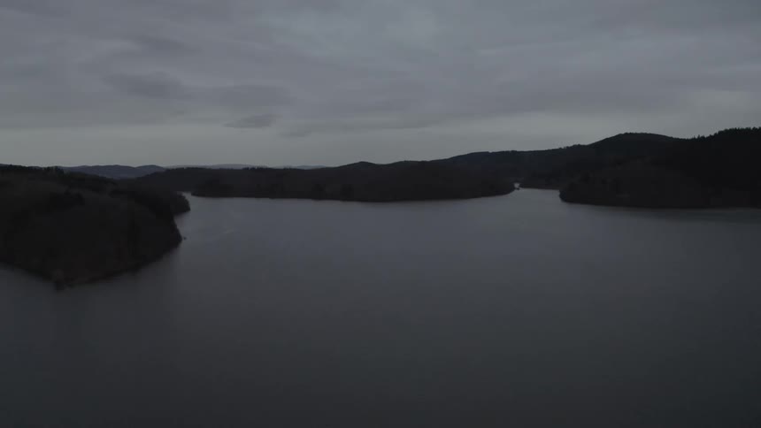 A static aerial view of a dark, serene lake surrounded by rolling hills under a gloomy, overcast sky at dusk