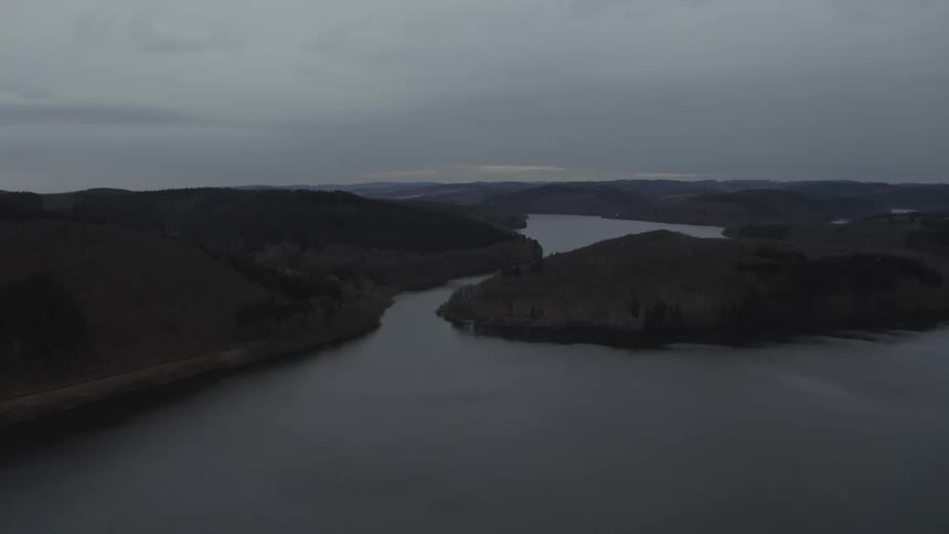 An aerial view shows a winding river flowing through dark, forested hills under a cloudy, twilight sky