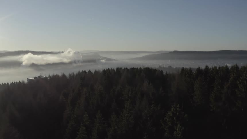 Aerial shot flying over a dark forest to reveal a misty valley with an industrial town in the background during sunrise