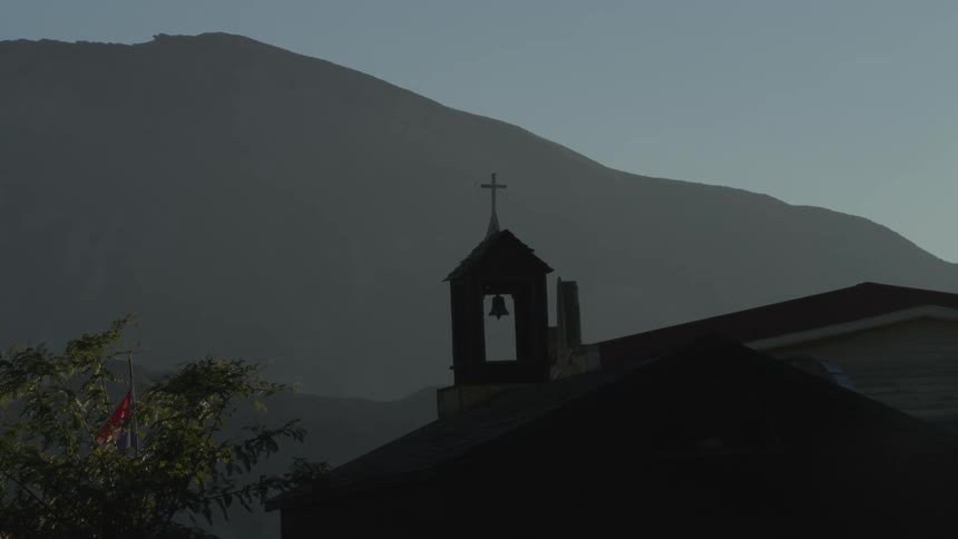 A serene view of a silhouetted church bell tower with a cross on top against a backdrop of hazy mountains at dawn