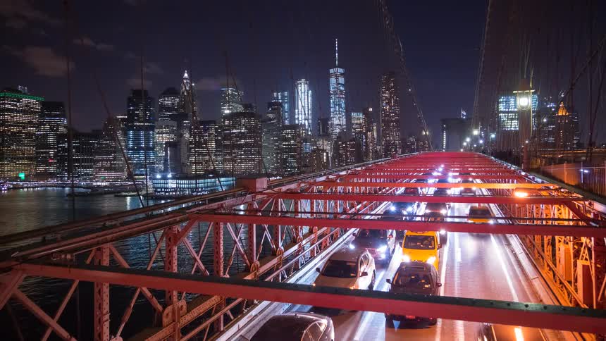 A nighttime timelapse captures the stream of traffic on the Brooklyn Bridge with the glowing Manhattan skyline as a backdrop