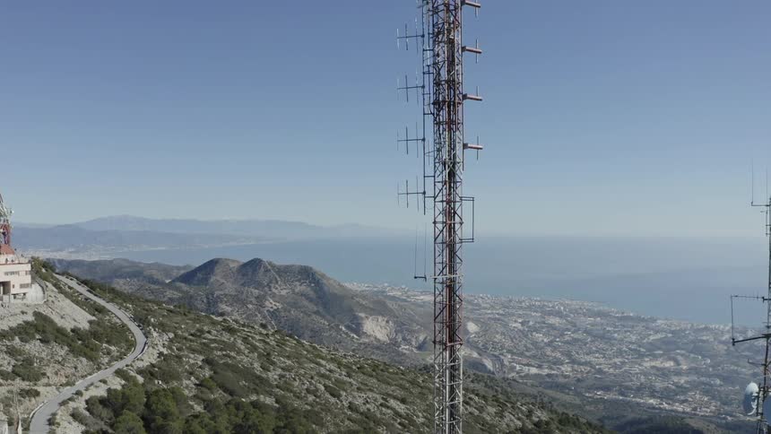 A large telecommunications tower stands on a mountaintop overlooking a coastal city and the sea under a clear blue sky