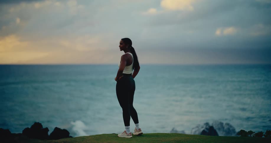 A fit woman in athletic wear stands on a grassy cliff, watching a powerful ocean wave crash in dramatic slow motion at dusk