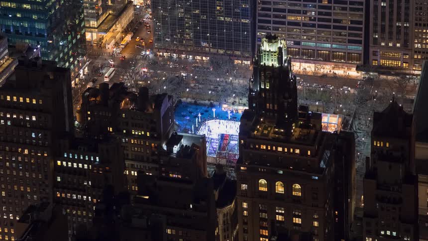 High-angle time-lapse of the brightly lit ice skating rink at Bryant Park in New York City at night, surrounded by towering skyscrapers and busy traffic