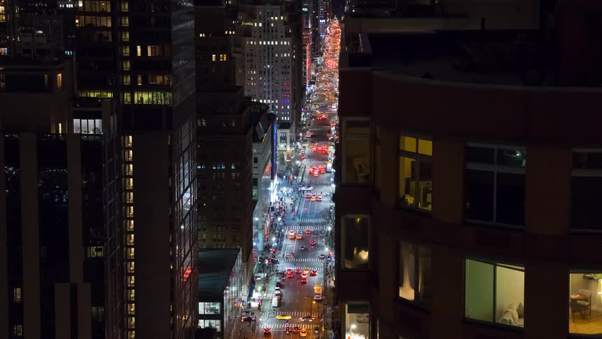 A high-angle time-lapse of a bustling New York City street at night with streaks of light from heavy traffic between towering skyscrapers