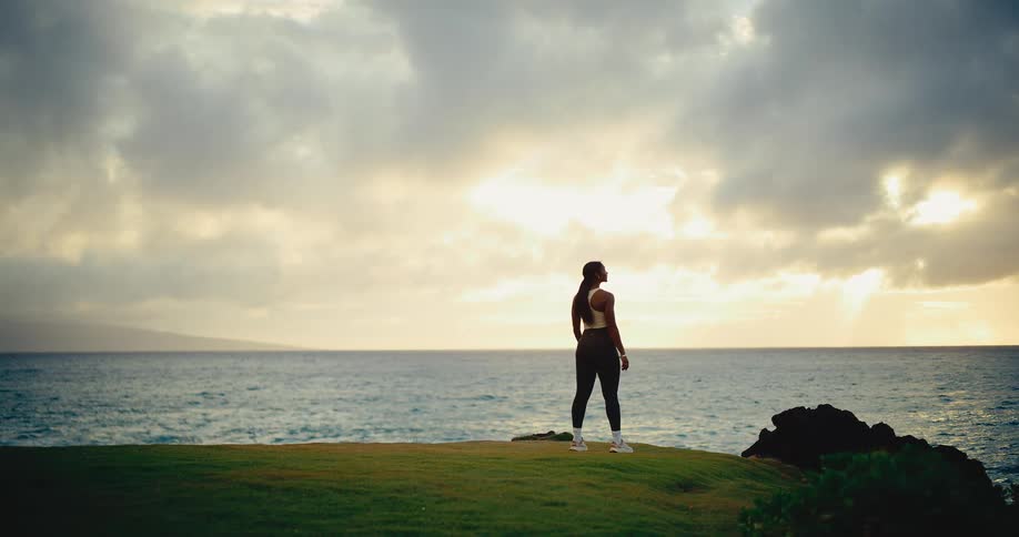 A beautiful athletic woman stands peacefully on a green cliff overlooking the ocean at sunrise, a moment of reflection and strength
