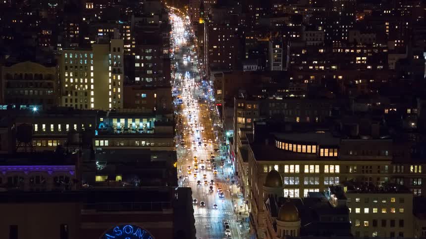 A vibrant time-lapse captures the energy of a bustling city avenue at night, with streams of traffic flowing between illuminated buildings