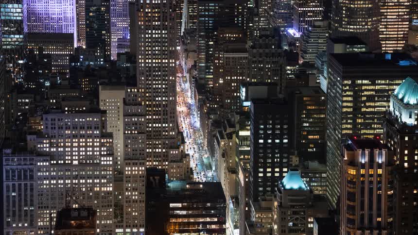 An aerial timelapse of traffic moving down a bustling New York City street surrounded by illuminated skyscrapers at night