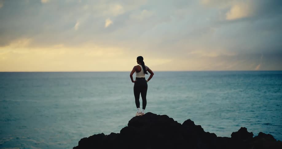 An athletic woman stands on a rocky precipice, gazing out at the vast ocean during a dramatic and cloudy sunset