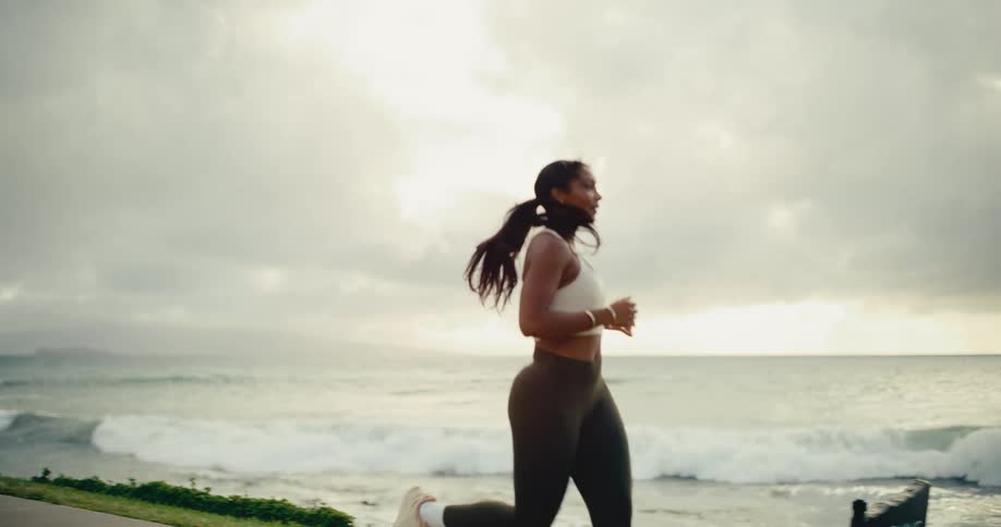 A fit woman jogs in slow motion along a scenic coastal path with the ocean waves crashing in the background