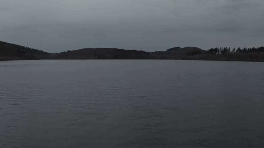 A slow aerial pullback reveals a vast, dark lake surrounded by forested hills on a calm and moody overcast evening