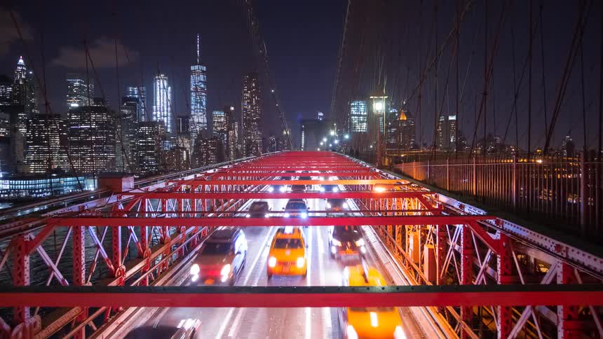 A dynamic night time-lapse from the Brooklyn Bridge captures the constant flow of traffic with the glittering Manhattan skyline in the background