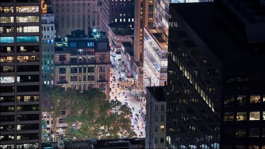 A high-angle time-lapse of bustling city traffic at night, framed by towering skyscrapers with illuminated windows