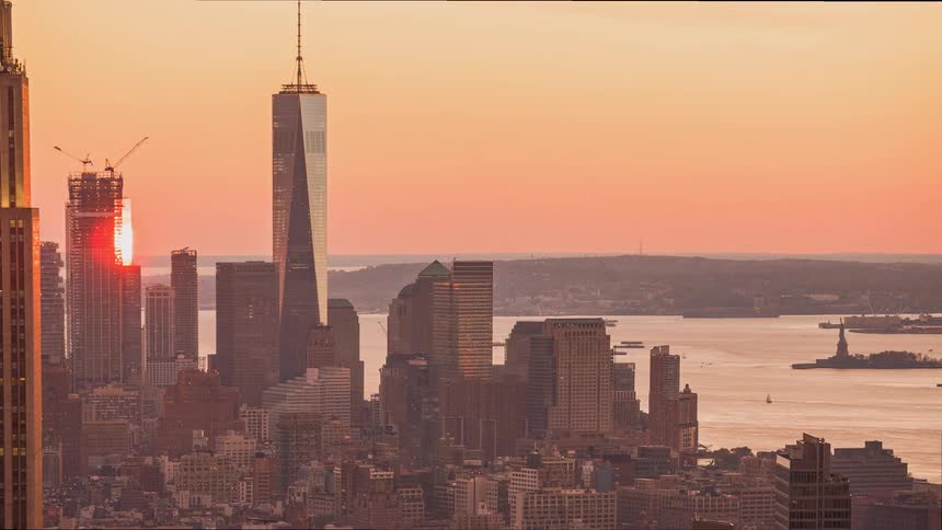A scenic time-lapse of the sun setting behind the New York City skyline, casting a warm glow over the Freedom Tower and the Statue of Liberty