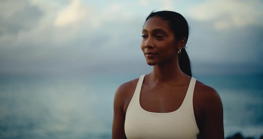 An athletic young woman in workout clothes peacefully gazes at the ocean horizon during a beautiful, cloudy sunset