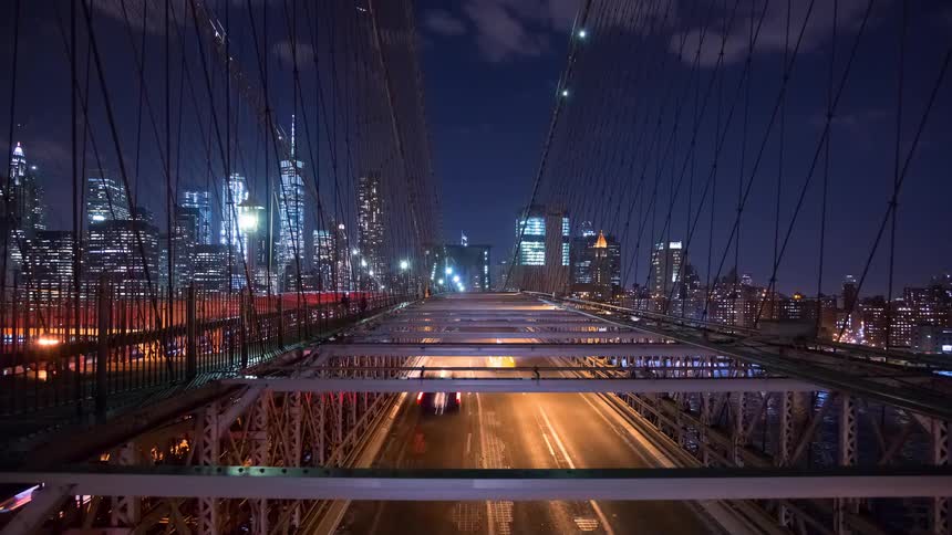 A dynamic time-lapse captures streaks of light from traffic crossing the Brooklyn Bridge at night, with the iconic Manhattan skyline glowing in the background