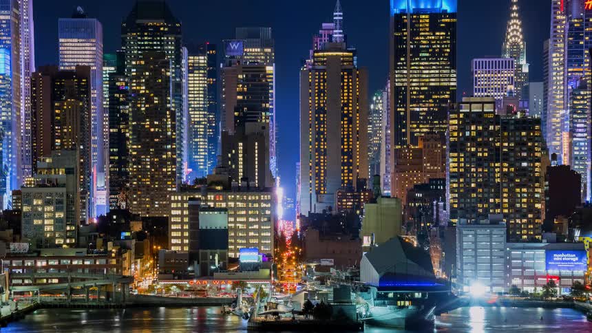 A dazzling night timelapse captures the vibrant energy of the New York City skyline, with illuminated skyscrapers and bustling traffic reflected in the water
