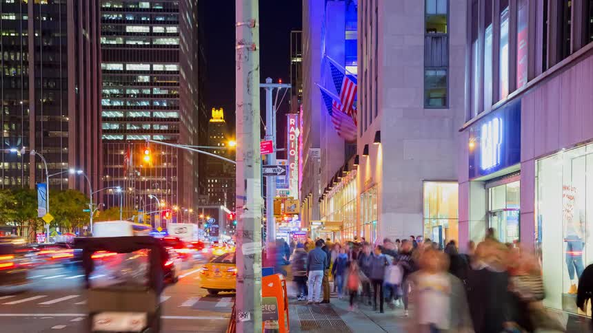 A vibrant time-lapse captures the bustling energy of a New York City street corner near Radio City Music Hall at night, with streams of traffic and crowds of pedestrians
