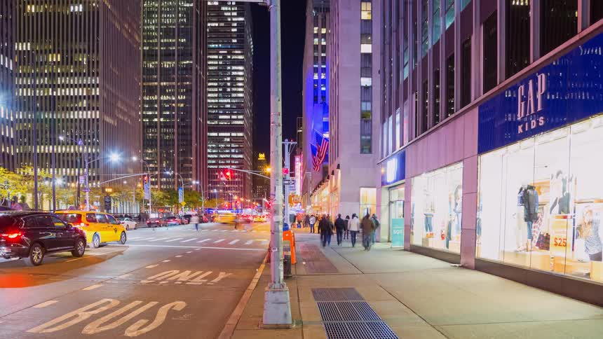 A nighttime time-lapse of a bustling New York City street corner shows the blurred motion of pedestrians and traffic