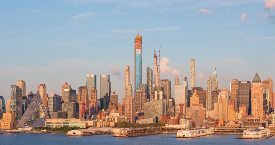 The Manhattan skyline glows in the warm light of sunset as clouds drift across the sky and a boat sails on the Hudson River