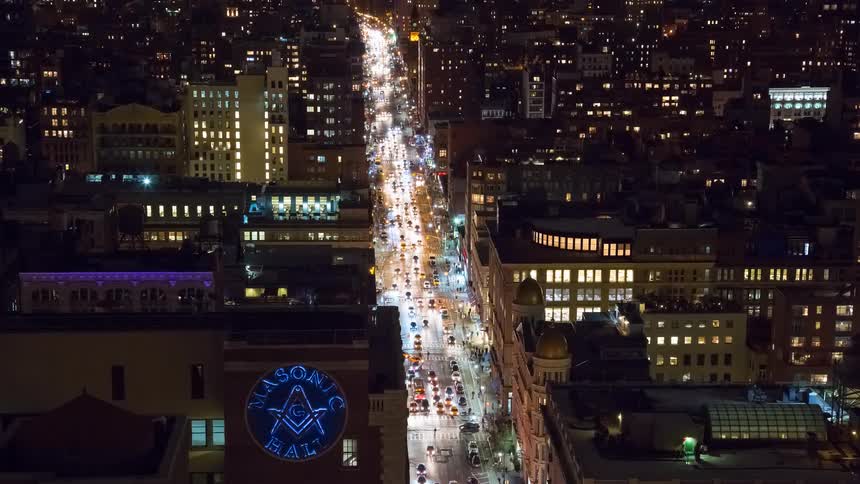 A high-angle time-lapse captures the bustling nightlife of a city street, with traffic lights streaking past the illuminated Masonic Hall