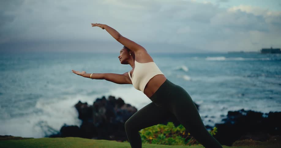 Graceful and athletic woman practices a serene yoga flow on a grassy cliff with powerful waves crashing on the rocks below