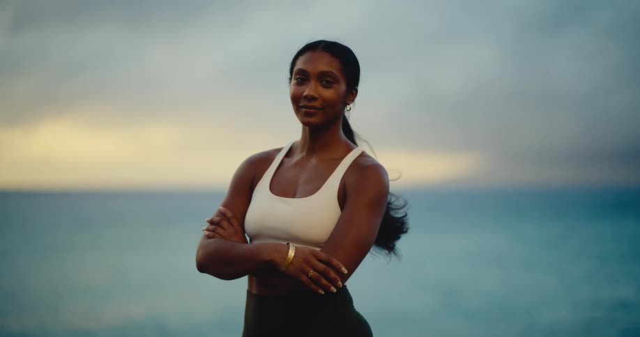 A confident and beautiful athletic woman looks directly into the camera with the ocean behind her at sunset in slow motion