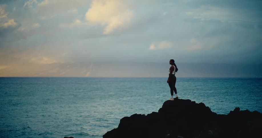 A woman in athletic wear stands on a rocky outcropping, peacefully looking out at the ocean under a dramatic, cloudy sky