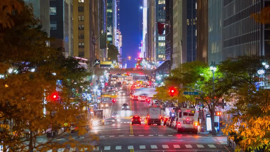 A bustling night-time timelapse captures the flow of traffic down a New York City avenue, surrounded by illuminated skyscrapers and framed by colorful autumn trees