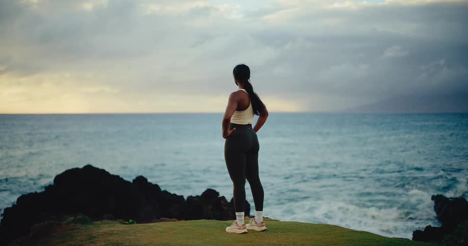 A fit woman in athletic wear stands on a grassy cliff, peacefully watching the ocean waves at sunrise